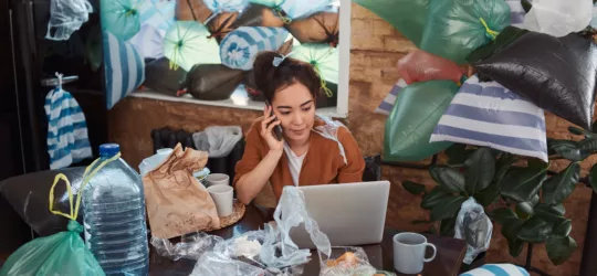 Young female communicating by phone in front of a laptop at her messy and cluttered apartment with rubbish everywhere