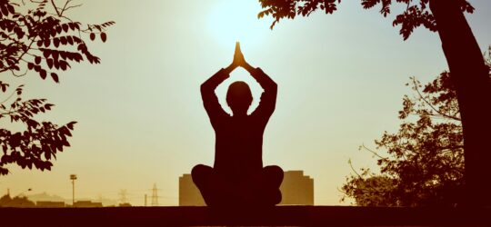 Person sitting cross-legged outdoors performing a yoga pose at sunrise or sunset, framed by tree branches- Junk-A-Haulics